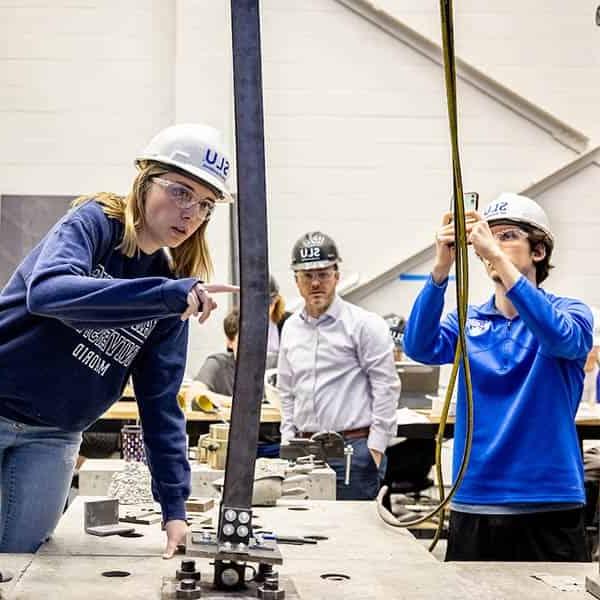 A female student wearing a hard hat points to a bend in a beam in a lab. Another male student, also wearing a hard hat, takes photos of the beam on a cell phone. A male instructor can be seen behind them watching them work.